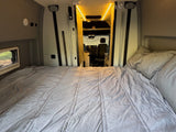 Cozy RV rental interior with a bed in the foreground and the driver's cabin visible. Bright lights illuminate the ceiling.