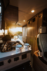 Warm sun streams into a cozy RV rental kitchen with a stove, French press, and "To Tour Shore" mugs.