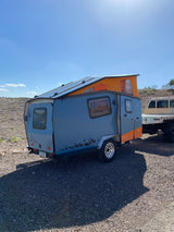 Unique gray & orange rv rental camper trailer with solar panels & pop-top, ready for outdoor adventure on rocky ground.
