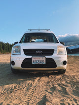 White Ford Transit Connect RV rental on a dirt road by a lake and forest. Ideal for camping and road trip adventures.