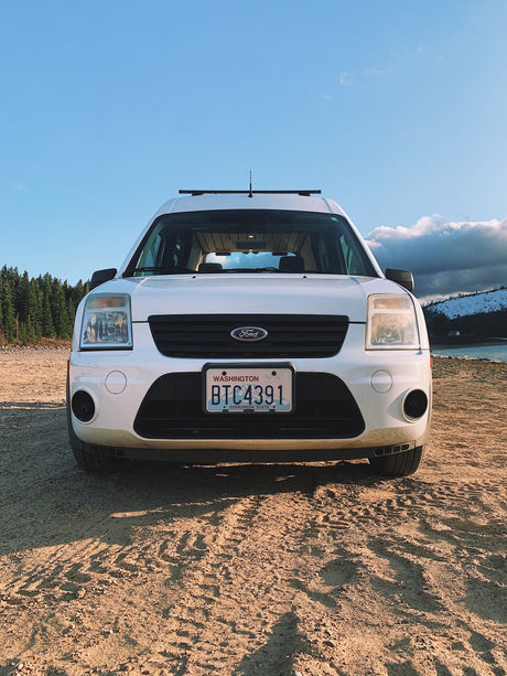 White Ford Transit Connect RV rental on a dirt road by a lake and forest. Ideal for camping and road trip adventures.
