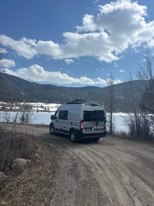 Grey RV rental on a dirt road by a serene, partially frozen lake, framed by snowy mountains and a cloudy blue sky.