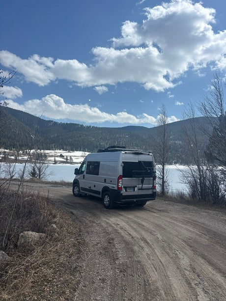 Grey RV rental on a dirt road by a serene, partially frozen lake, framed by snowy mountains and a cloudy blue sky.