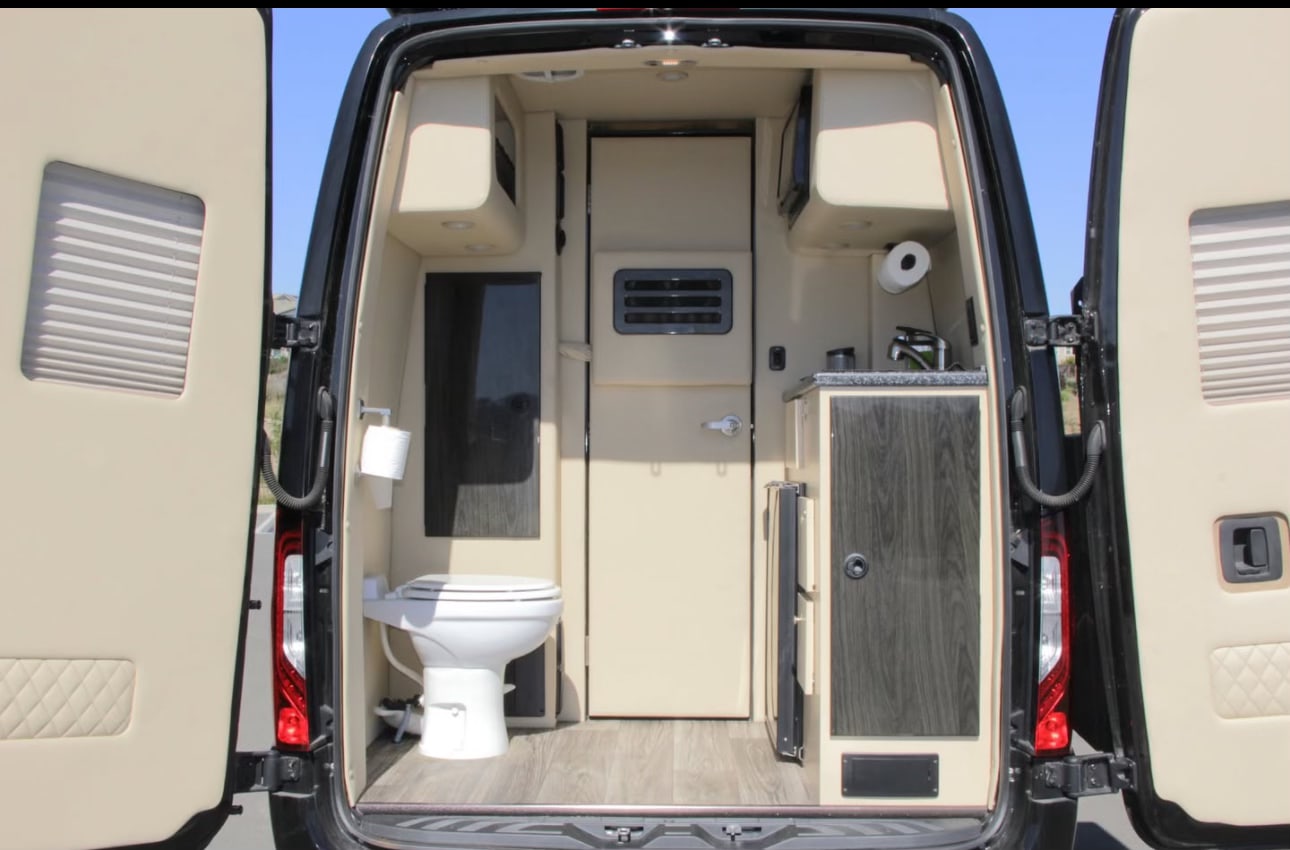 **RV rental**'s rear interior with open doors reveals a toilet, sink, and compact kitchen. Beige and dark wood design. — photo 10