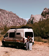 Explore with an **rv rental**! Two women pose by a Storyteller Overland camper van amidst rugged mountains and trees.