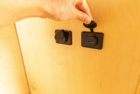 Hand opens a weather-sealed USB-A/C charging port in an RV rental's wooden interior, offering convenient device power.