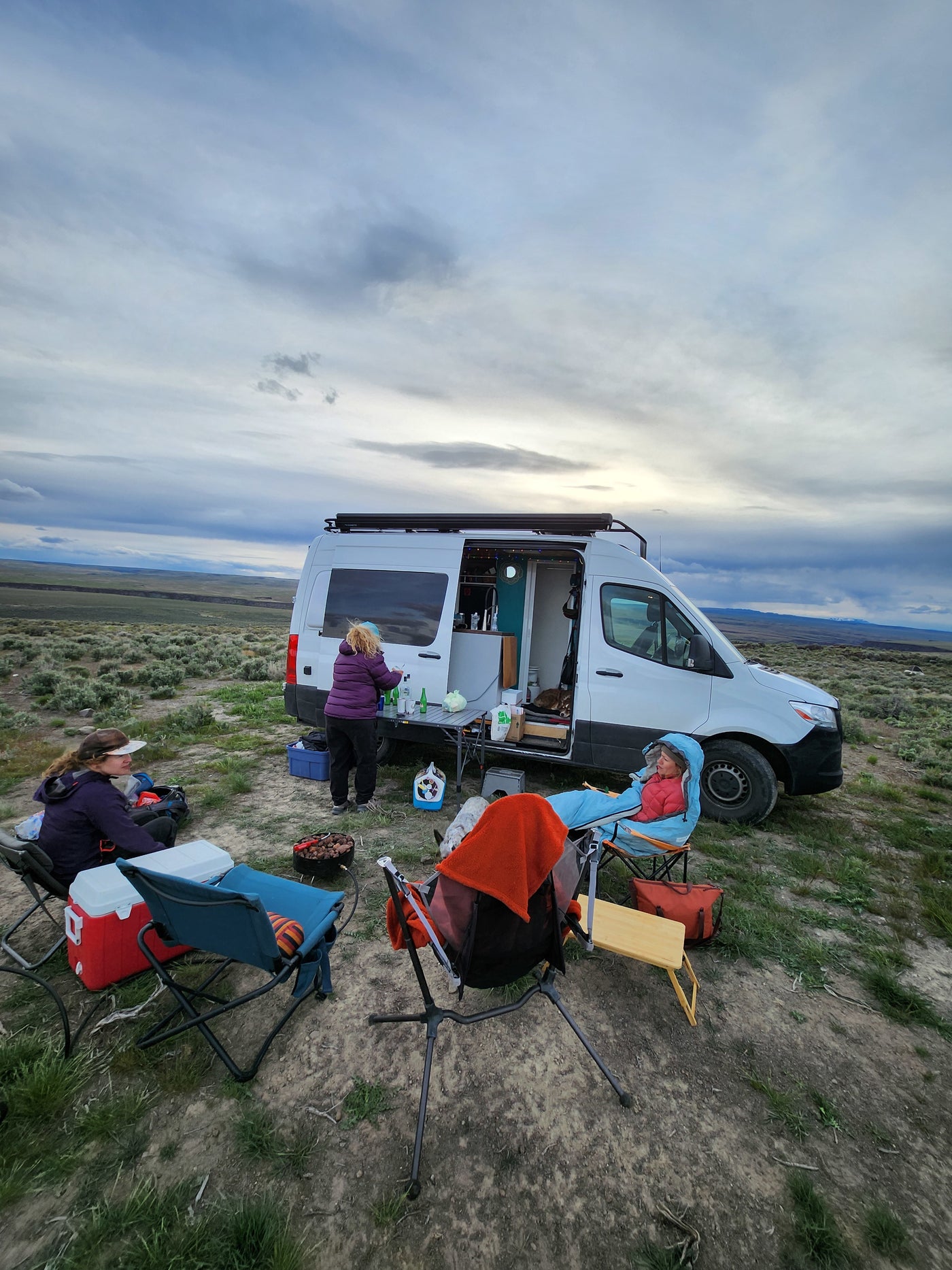 RV rental journey: people camping & cooking outside their camper van in a serene, open landscape under a cloudy sky. — photo 6