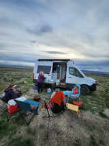 RV rental journey: people camping & cooking outside their camper van in a serene, open landscape under a cloudy sky.