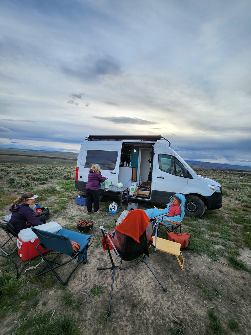 RV rental journey: people camping & cooking outside their camper van in a serene, open landscape under a cloudy sky. — photo 6