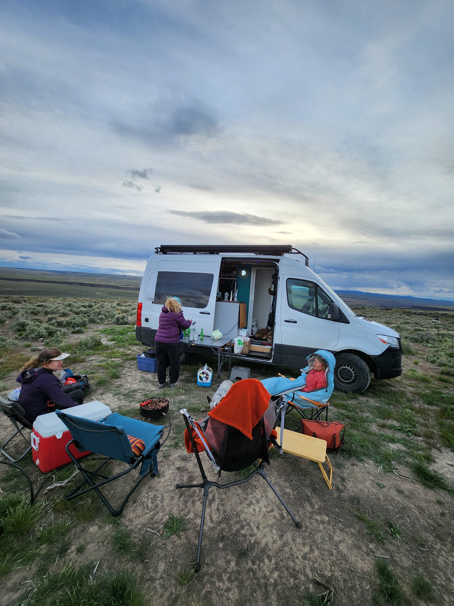 RV rental journey: people camping & cooking outside their camper van in a serene, open landscape under a cloudy sky.