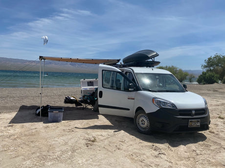 RV rental van set up on a sandy beach. Awning extended, camping gear, and a lake view with kitesurfer and mountains. Perfect for adventure.