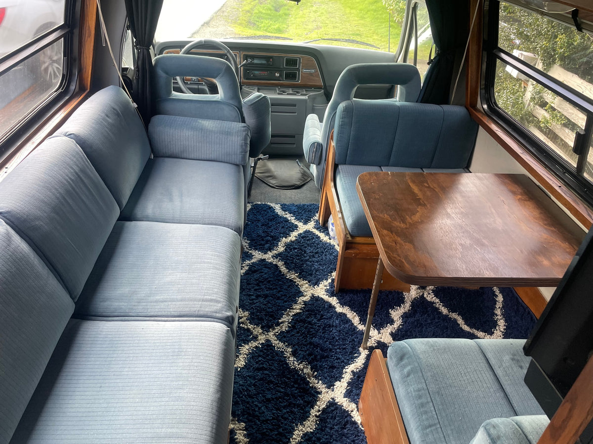 Light blue ribbed seating lines this RV rental's interior, featuring a dark geometric rug, wooden table, and front cockpit. — photo 5