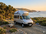 RV rental camper van parked on a scenic coastal road at sunset, overlooking the Pacific Ocean and hills. Perfect for a road trip!