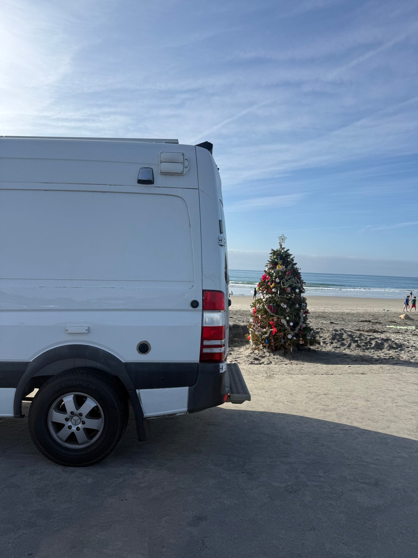 A white rv rental van parked on a sandy beach next to a festive Christmas tree, with the ocean in the background.