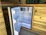 RV rental: Bright, open refrigerator with empty shelves in a custom wood RV interior, showing cabinets, drawers, and a bed.