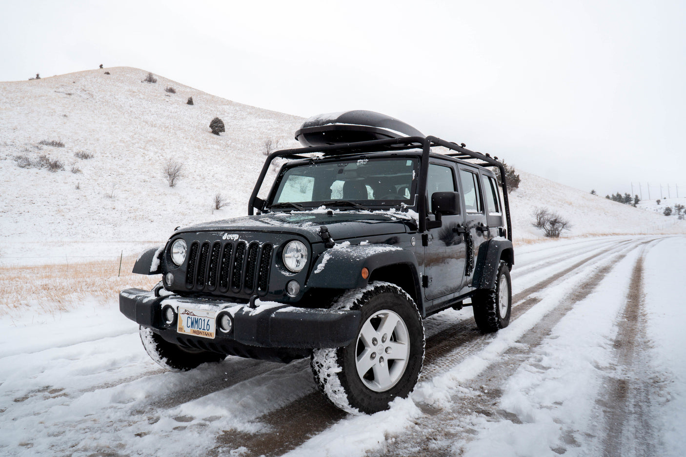 Dark green Jeep Wrangler with roof cargo box on a snowy winter road. Perfect for off-road adventure or an RV rental trip.