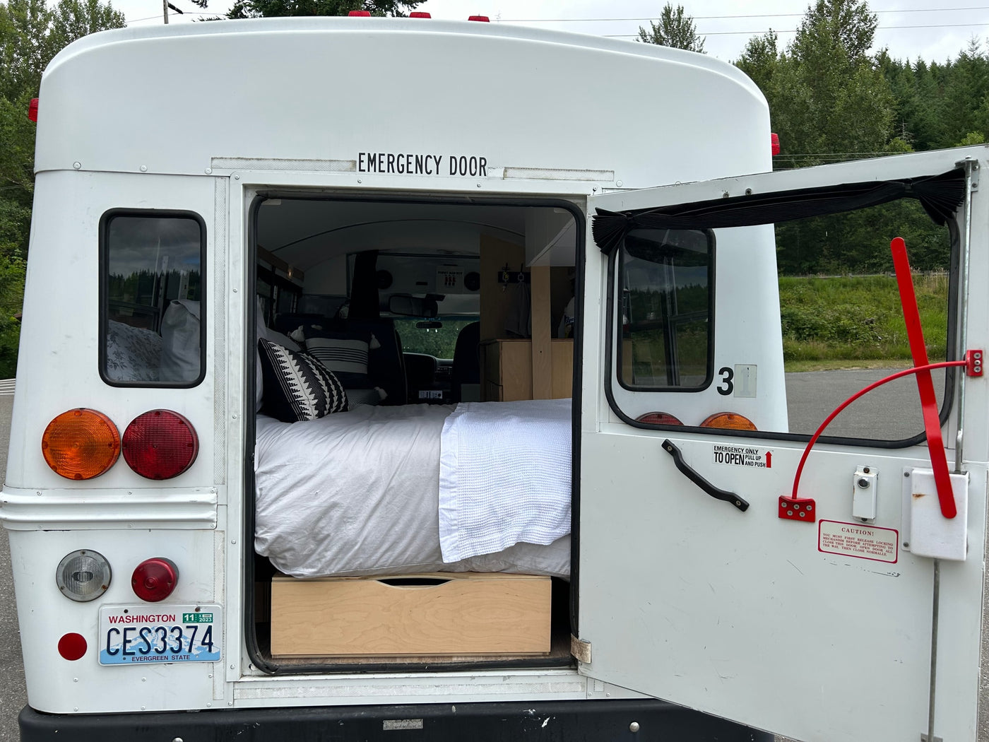 Rear of a white bus conversion rv rental with its emergency door open, revealing a cozy bed and storage within. WA plate visible. — photo 9