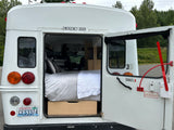 Rear of a white bus conversion rv rental with its emergency door open, revealing a cozy bed and storage within. WA plate visible.