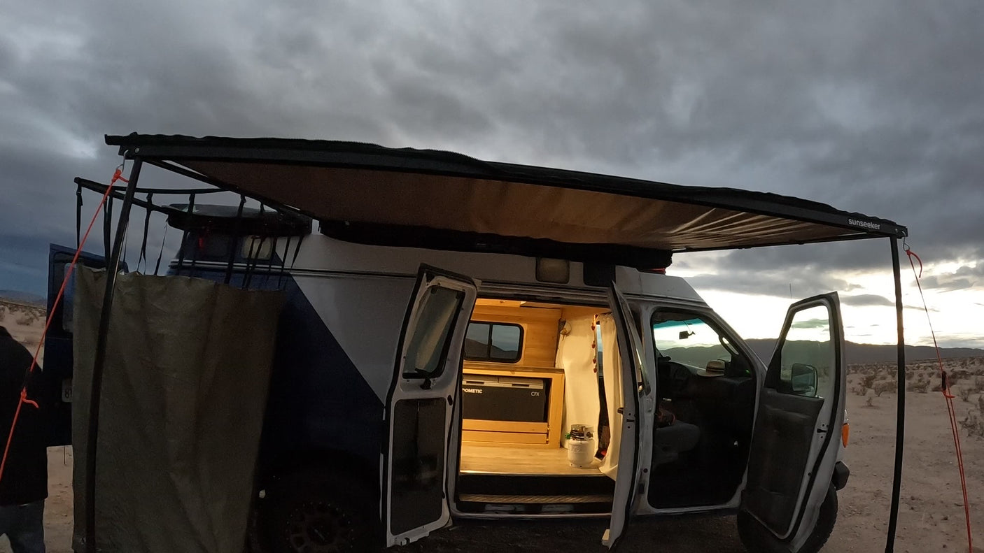 Custom RV rental camper van with extended awning. Open doors reveal cozy wooden interior & fridge in a desert landscape.