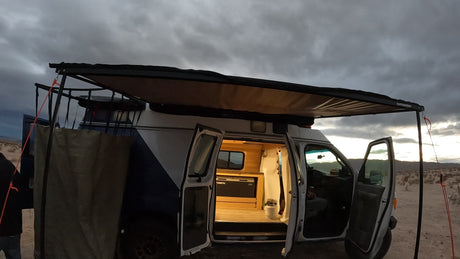 Custom RV rental camper van with extended awning. Open doors reveal cozy wooden interior & fridge in a desert landscape.