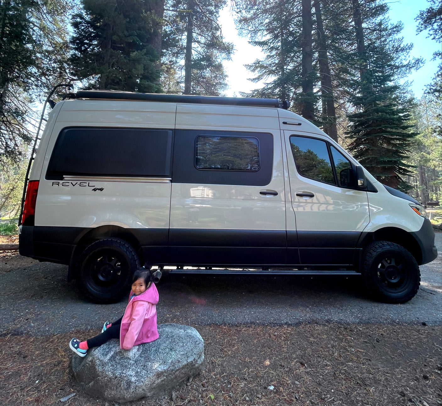 A child sits by a cream-colored Revel 4x4 camper van in a sunny forest, perfect for an RV rental adventure.