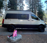 A child sits by a cream-colored Revel 4x4 camper van in a sunny forest, perfect for an RV rental adventure.