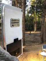 White RV rental door with a 'Happy Campers Live Here' sign, open to a peaceful forest setting with tall trees and dry ground.