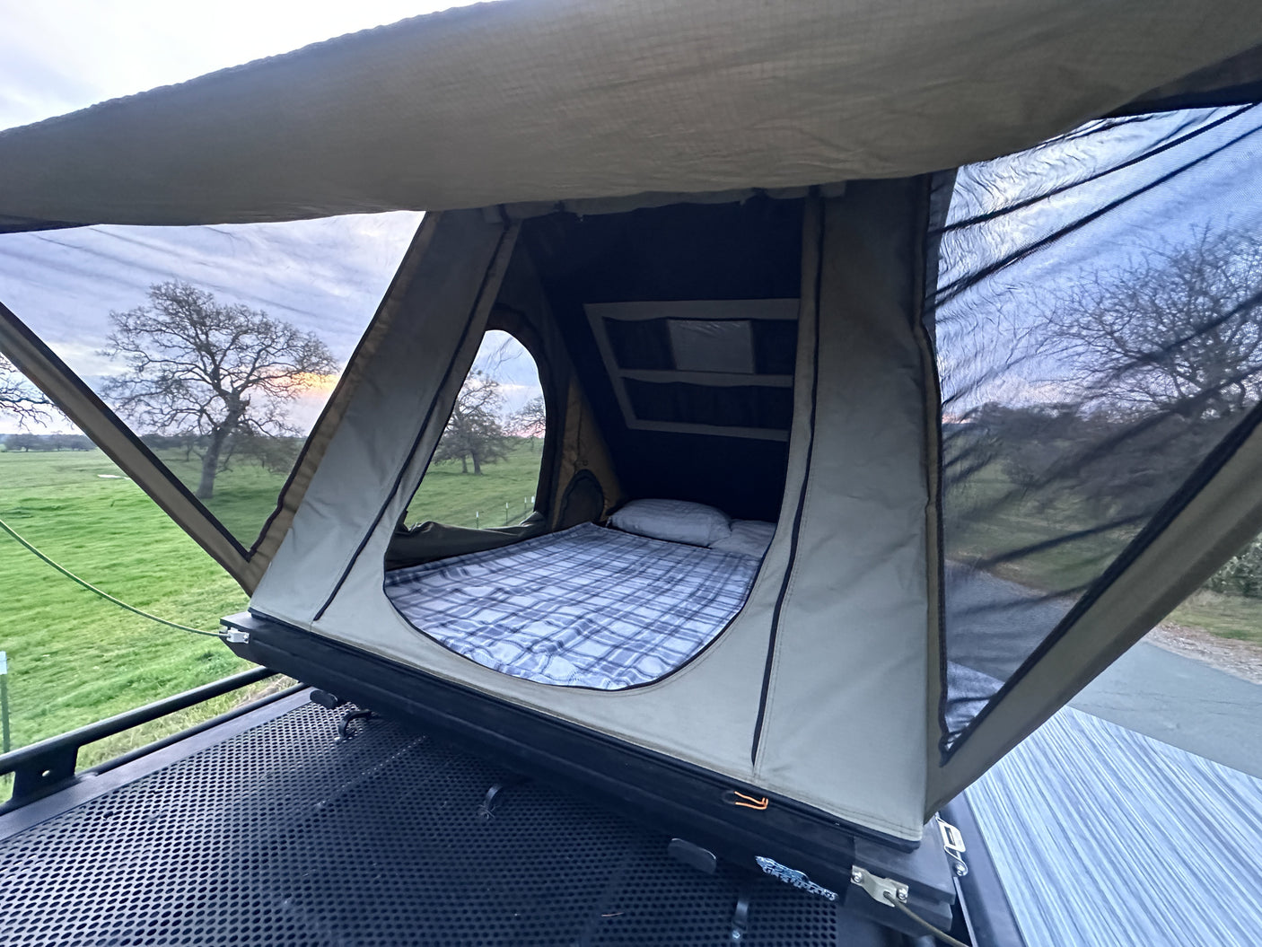 RV rental rooftop tent, ready for cozy nights with a comfy bed and views of lush fields and trees at dusk.