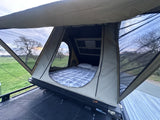 RV rental rooftop tent, ready for cozy nights with a comfy bed and views of lush fields and trees at dusk.