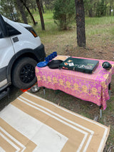 RV rental campsite with outdoor kitchen setup: propane stove, cast iron pan, camping dishes on a colorful table, and an outdoor mat.