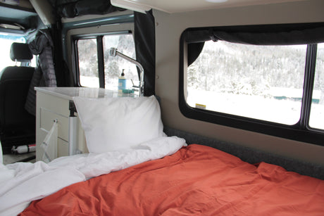 Cozy RV rental interior with a bed, small kitchen sink area, and scenic snowy landscape visible through the windows.