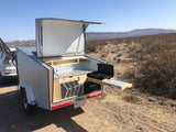 Rear view of a compact teardrop RV rental, open to reveal a full outdoor kitchen, sink, and stove in a desert setting.