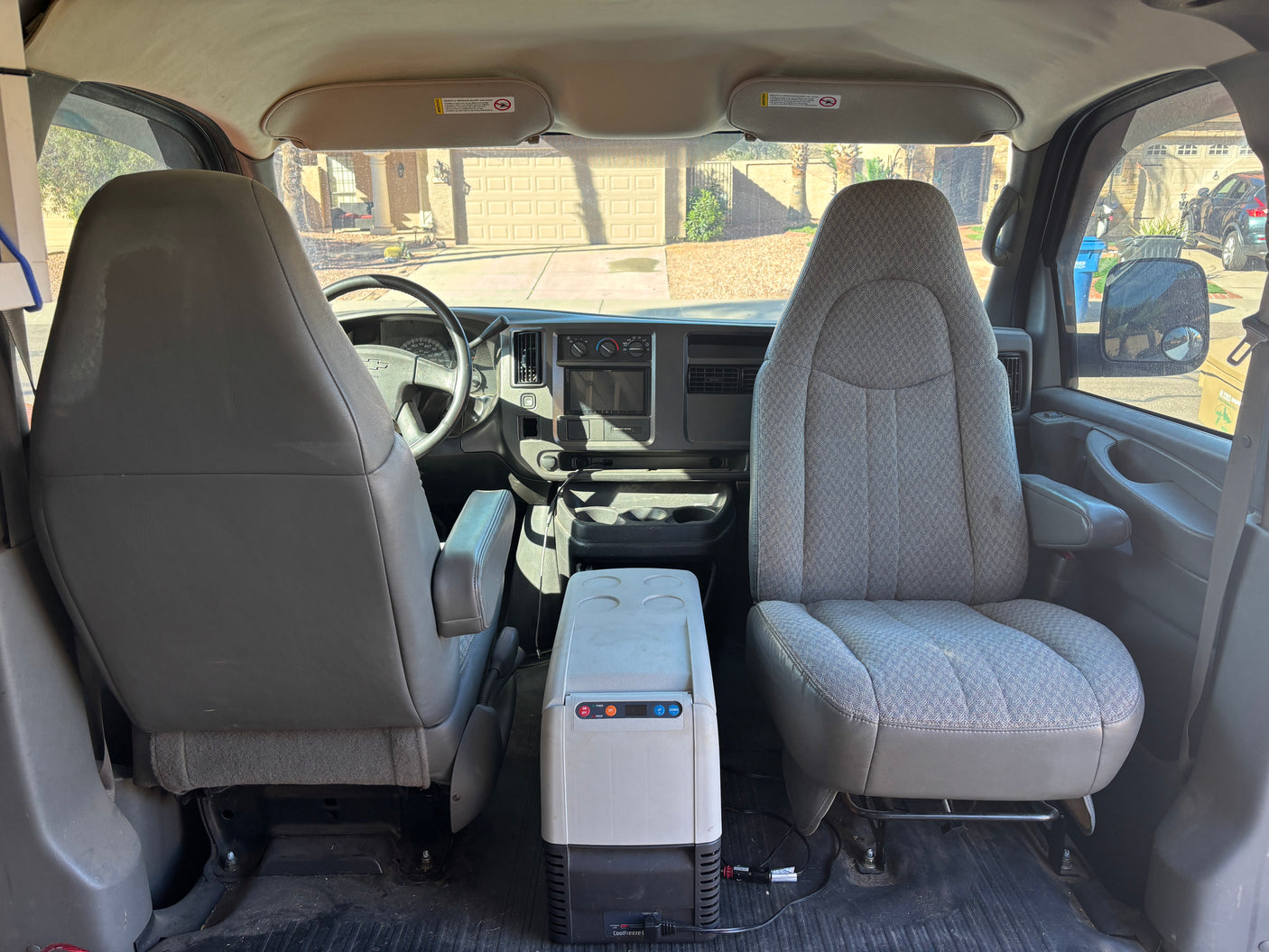 RV rental van's front cabin: driver/passenger seats, dashboard, and a portable fridge between them, with a house view outside.