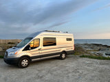 Silver campervan RV rental parked by a rocky ocean coast at sunset, reflecting colorful skies. Perfect for your next adventure.