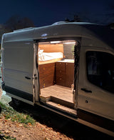 Night shot of a white converted camper van, perfect for rv rental. Its side door is open, revealing a cozy custom interior with a bed and cabinets.