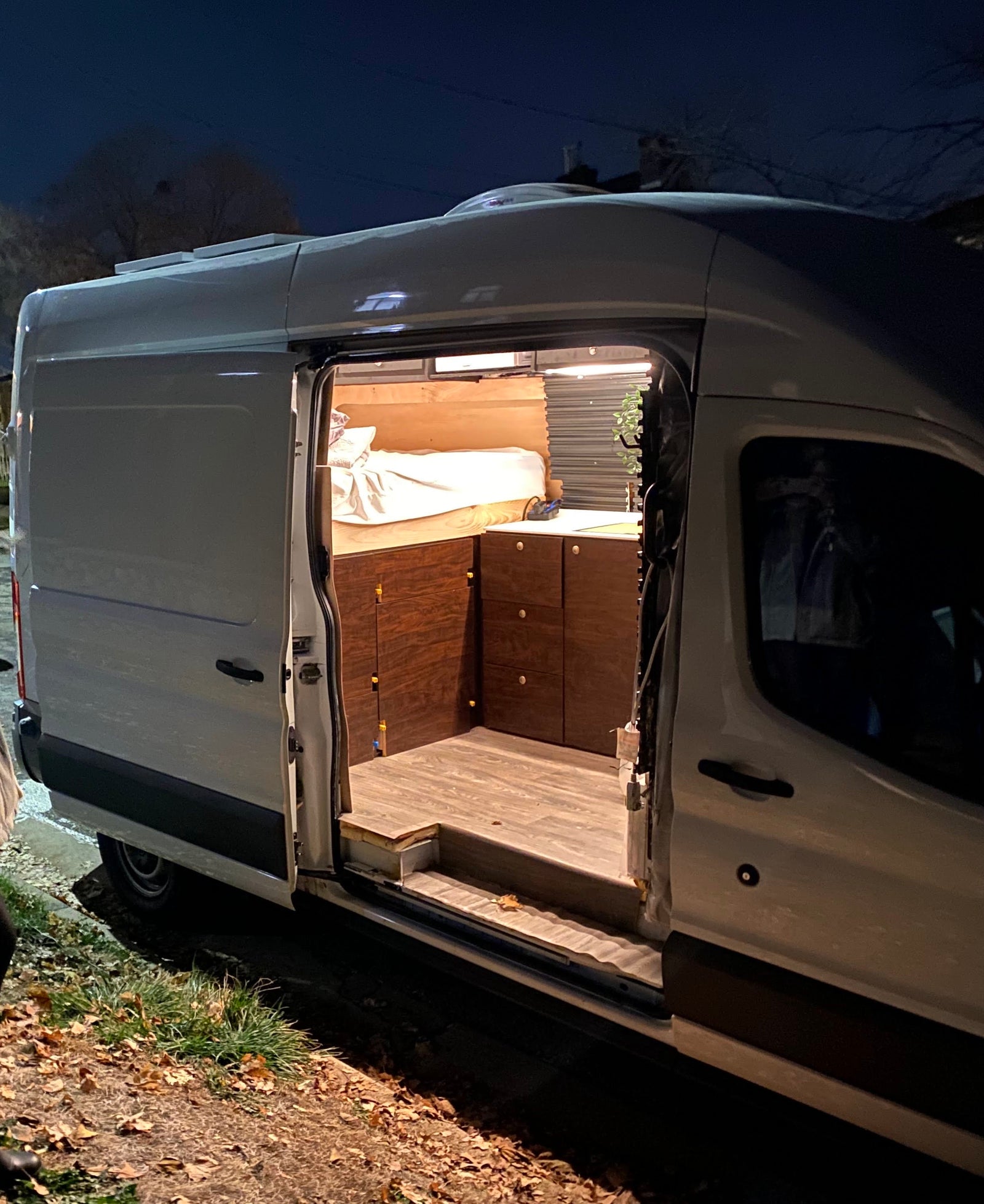 Night shot of a white converted camper van, perfect for rv rental. Its side door is open, revealing a cozy custom interior with a bed and cabinets.