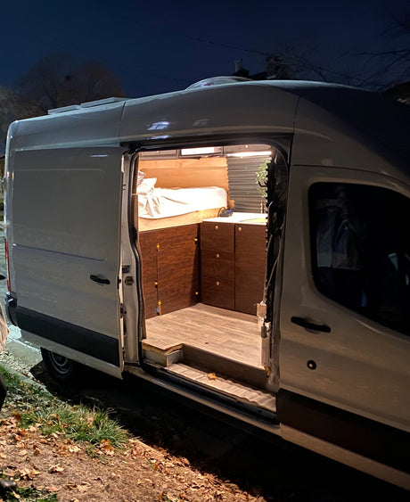 Night shot of a white converted camper van, perfect for rv rental. Its side door is open, revealing a cozy custom interior with a bed and cabinets.