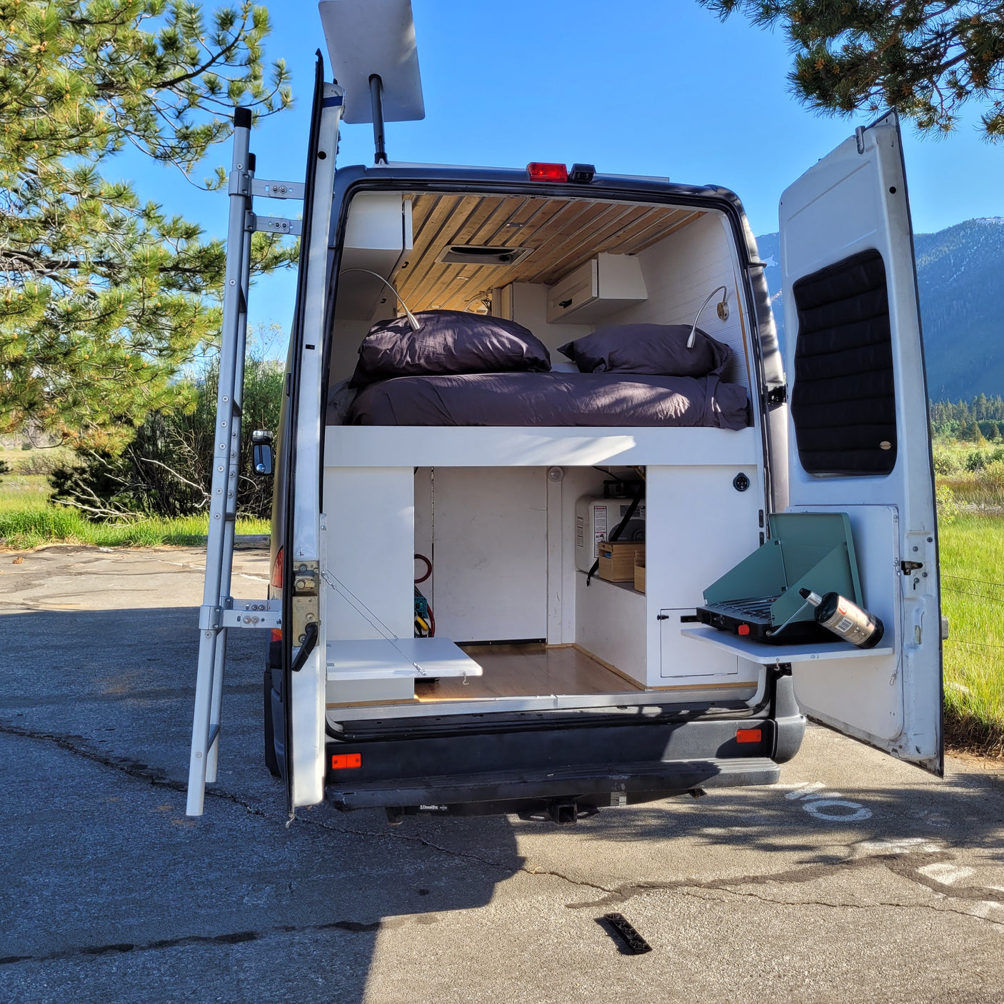This rv rental is equipped for adventure, featuring a comfortable interior bed, exterior ladder, and a convenient outdoor stove setup.