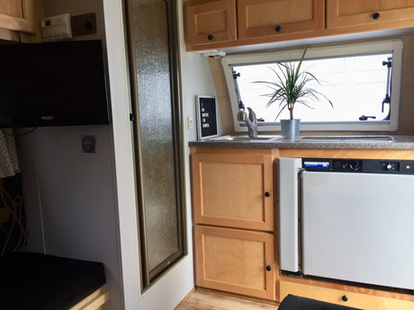 An rv rental interior shows a compact kitchen with a sink & plant, a mounted TV, and a frosted glass shower door.