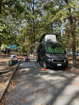 Dark gray pop-top rv rental camper van parked at a gravel campsite with a blue canopy & outdoor kitchen.