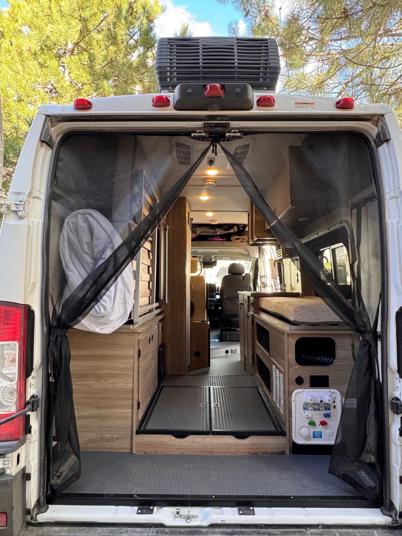 RV rental camper van interior: open rear doors reveal a bug screen, wood cabinetry, bed/seating area, and controls.