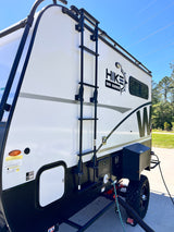 Adventure ready RV rental: Side view of a white Hike 100 Series travel trailer with ladder, utility hookups, and off-road tires.