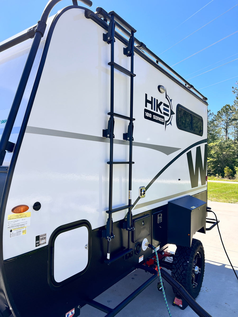  hitched to an SUV.|Side view of an RV rental at a campground. Power cable & blue coiled water hose are connected for setup.|Hike 100 Series RV rental with ladder & hookups visible