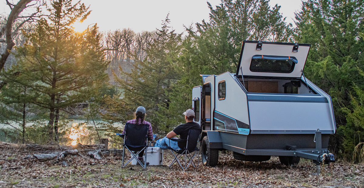A couple enjoys the outdoors by a lake, next to a small travel trailer. Consider an RV rental for your next camping trip.