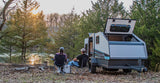 A couple enjoys the outdoors by a lake, next to a small travel trailer. Consider an RV rental for your next camping trip.