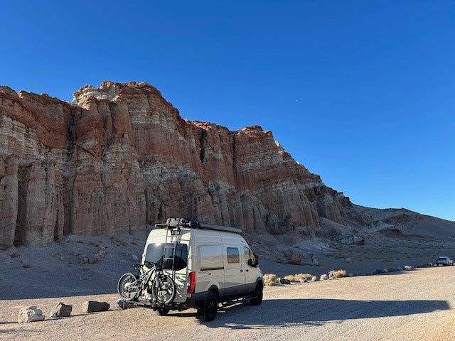 Exploring the desert: An RV rental van with bikes is parked before majestic red rock cliffs under a brilliant blue sky.