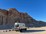 Exploring the desert: An RV rental van with bikes is parked before majestic red rock cliffs under a brilliant blue sky.