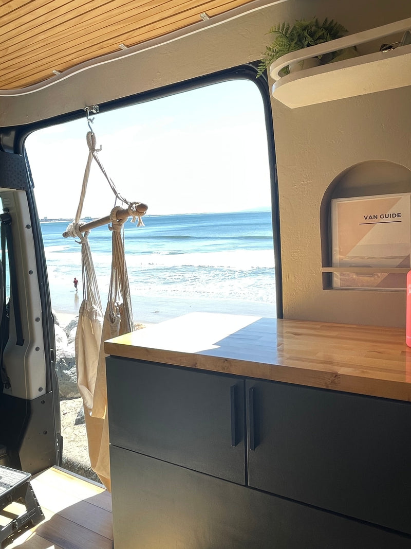 RV rental: Bright camper van interior with wooden ceiling & counter, custom storage, offering a clear beach and ocean view. — photo 8