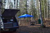 RV rental teardrop trailer at a fall campsite. Its open galley kitchen faces a blue canopy, picnic table, & fire pit.