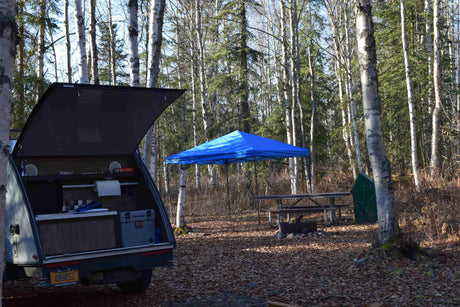 RV rental teardrop trailer at a fall campsite. Its open galley kitchen faces a blue canopy, picnic table, & fire pit.
